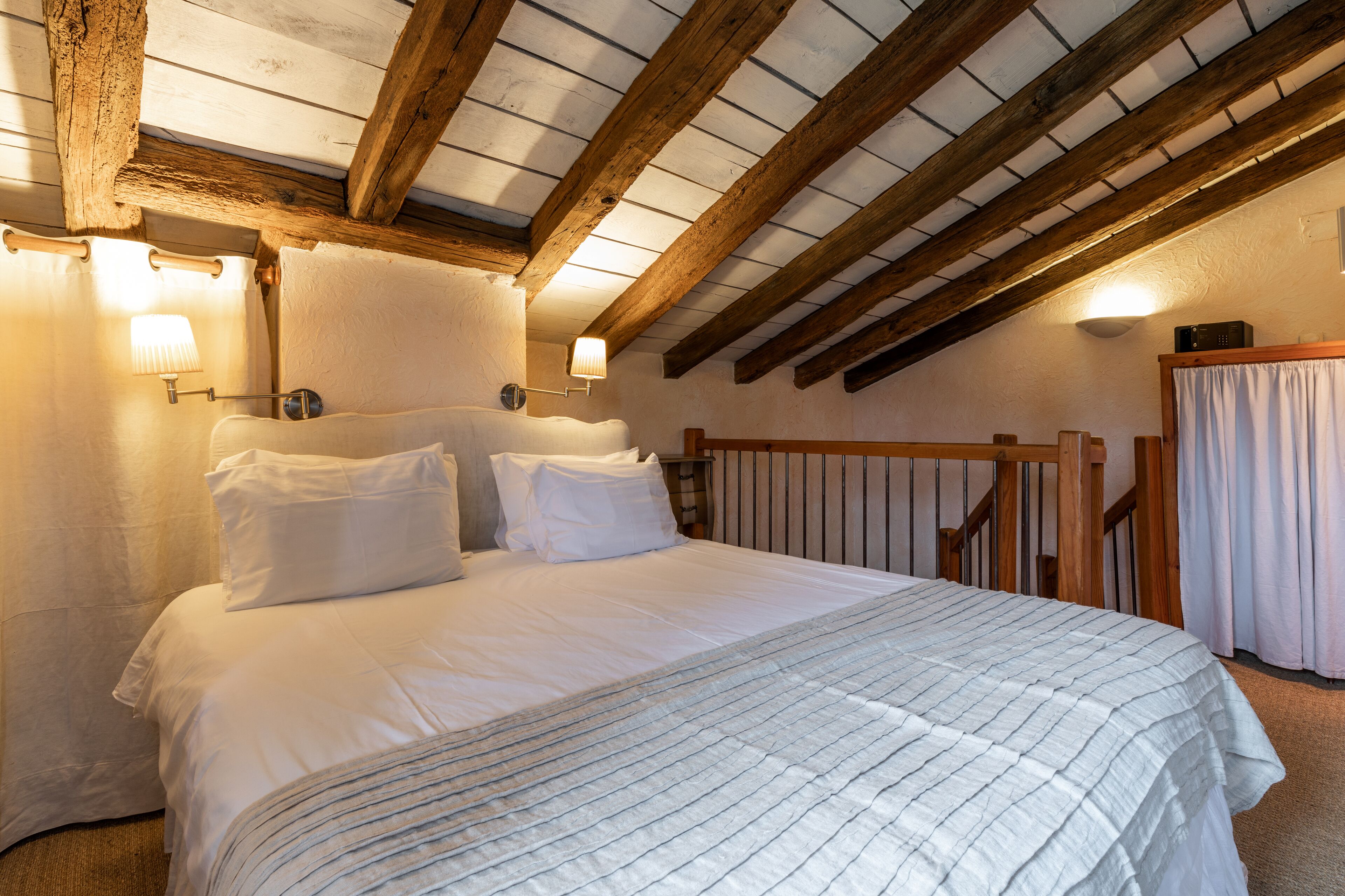 A bedroom with a large bed, wooden beams, and two wall-mounted lamps.