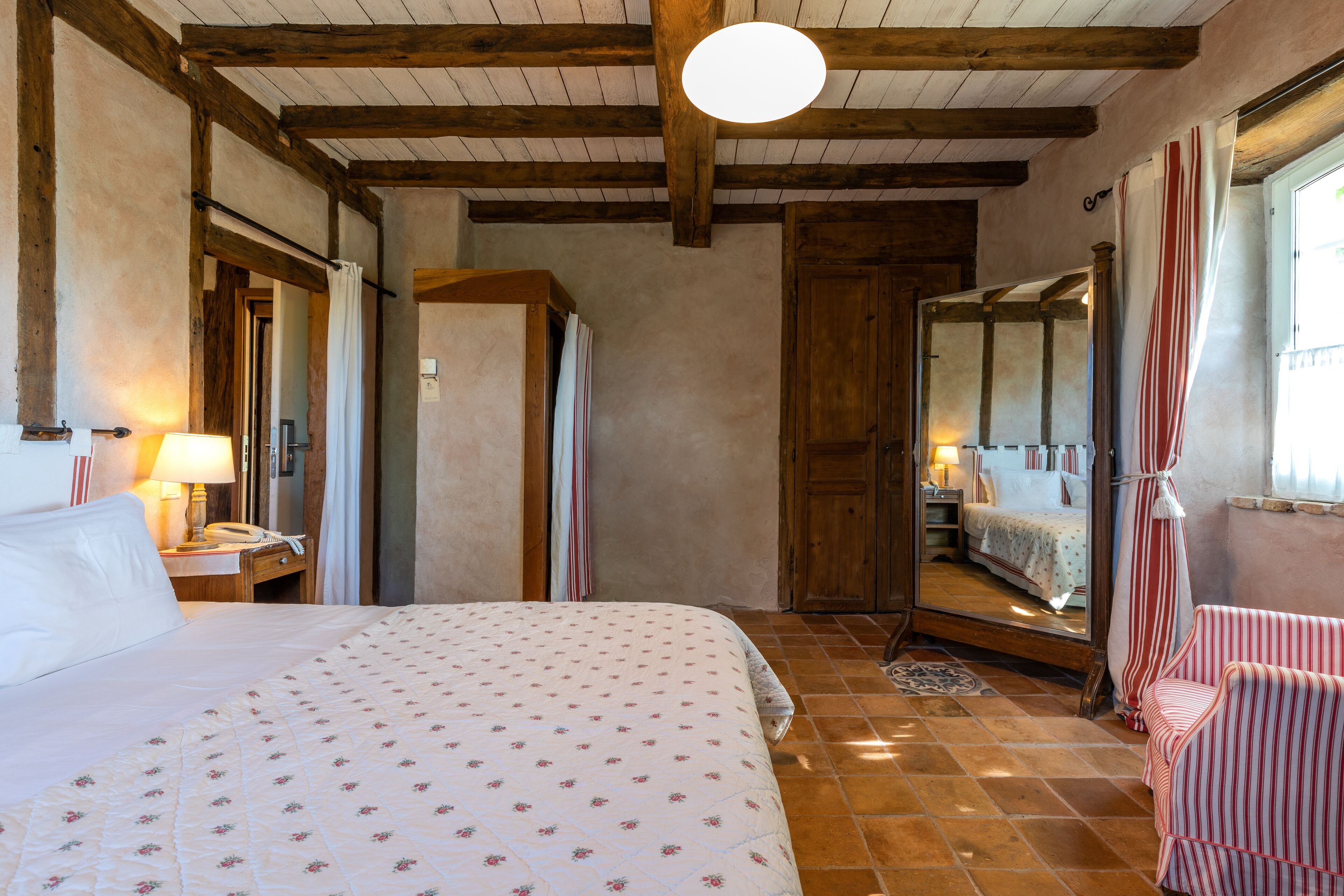 A bedroom with a large bed, wooden beams, a mirror, a chair, and a window with curtains.