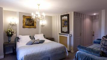 A hotel room with a large bed, a sitting area with a blue sofa, a chandelier, a wall-mounted mirror, and a vase with flowers.