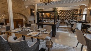 Un restaurante con techos altos, paredes de ladrillo visto y una araña. Las mesas están dispuestas con manteles blancos, cubiertos y copas de vino.