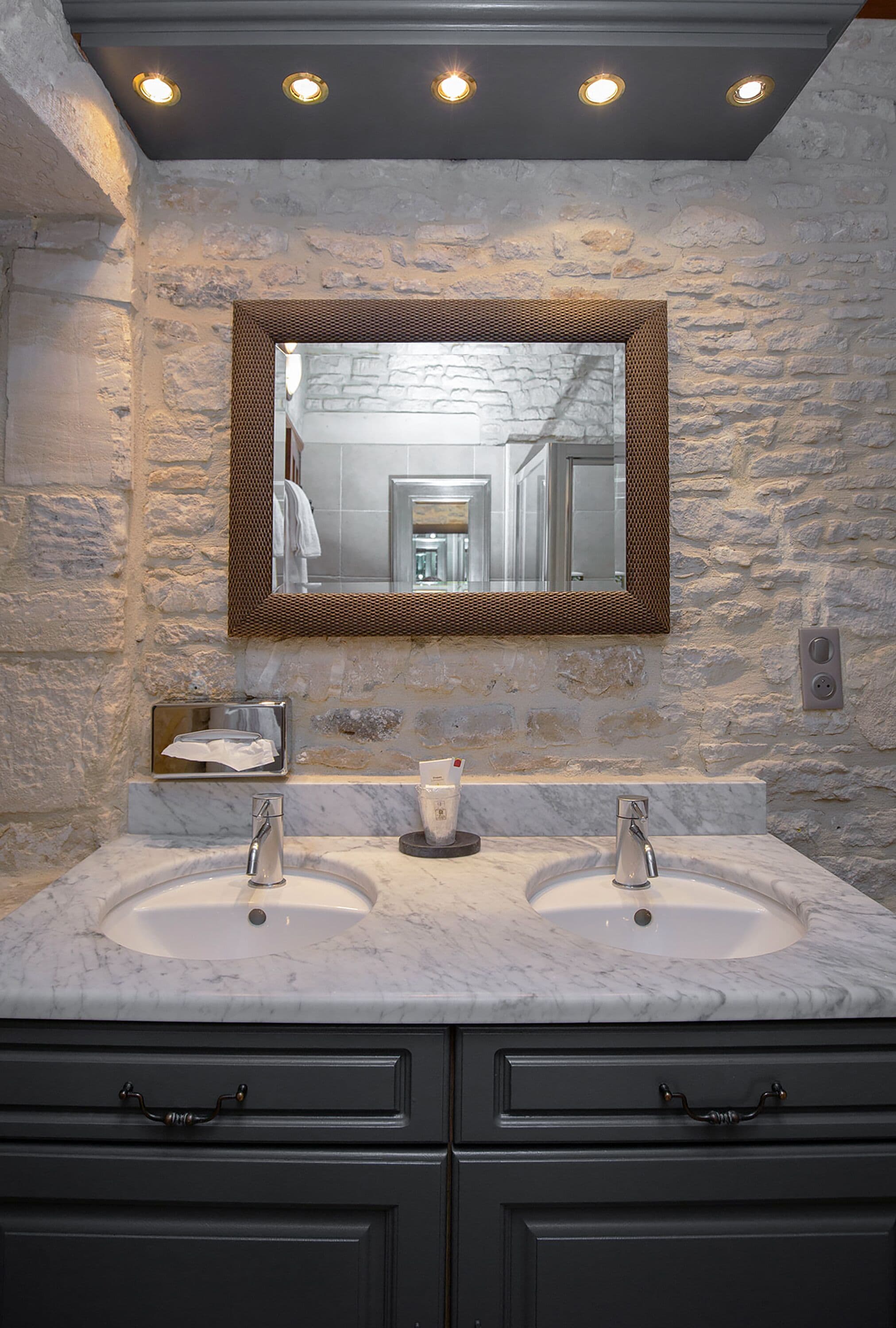 Un baño con pared de piedra, dos lavamanos, un espejo y un dispensador de toallas.