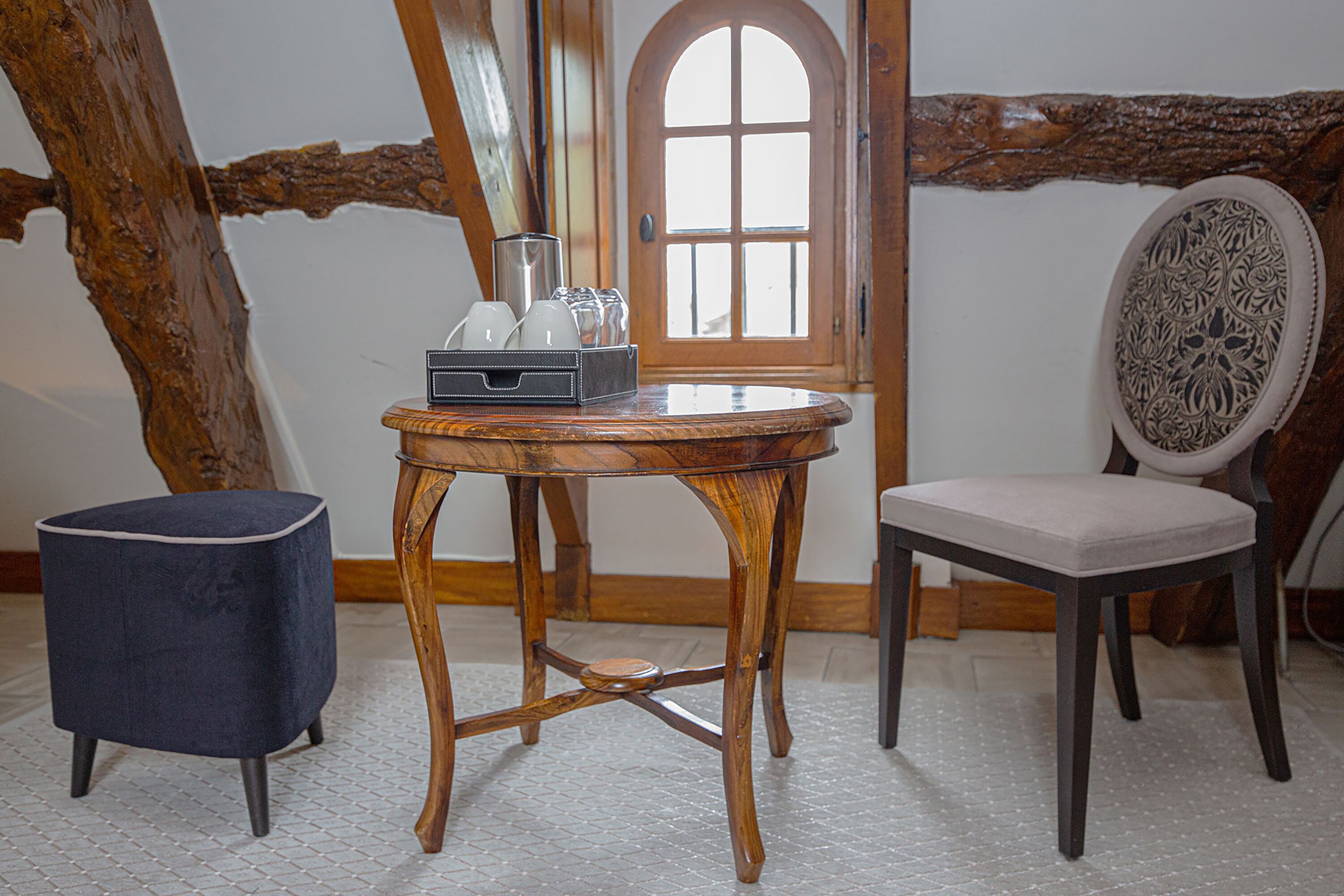 Une table en bois sur laquelle se trouve un plateau avec des verres et une carafe, un pouf bleu et une chaise avec un coussin à motifs, dans une pièce dont les poutres en bois sont apparentes et qui possède une porte en bois.