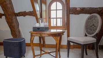 Une table en bois sur laquelle se trouve un plateau avec des verres et une carafe, un pouf bleu et une chaise avec un coussin à motifs, dans une pièce dont les poutres en bois sont apparentes et qui possède une porte en bois.