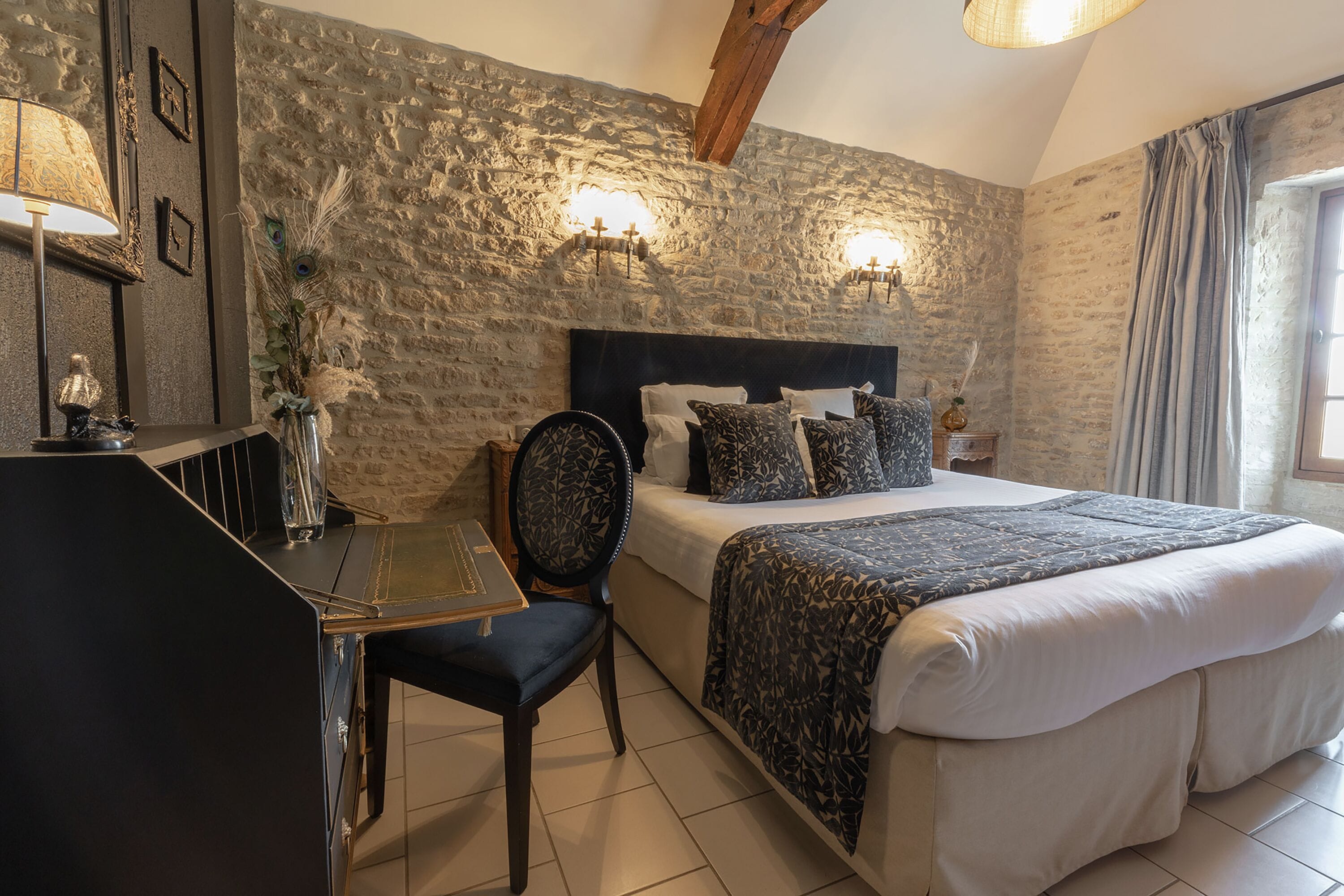 Un dormitorio con pared de piedra, una cama con ropa de cama estampada, un escritorio con una silla y dos lámparas fijadas a la pared.