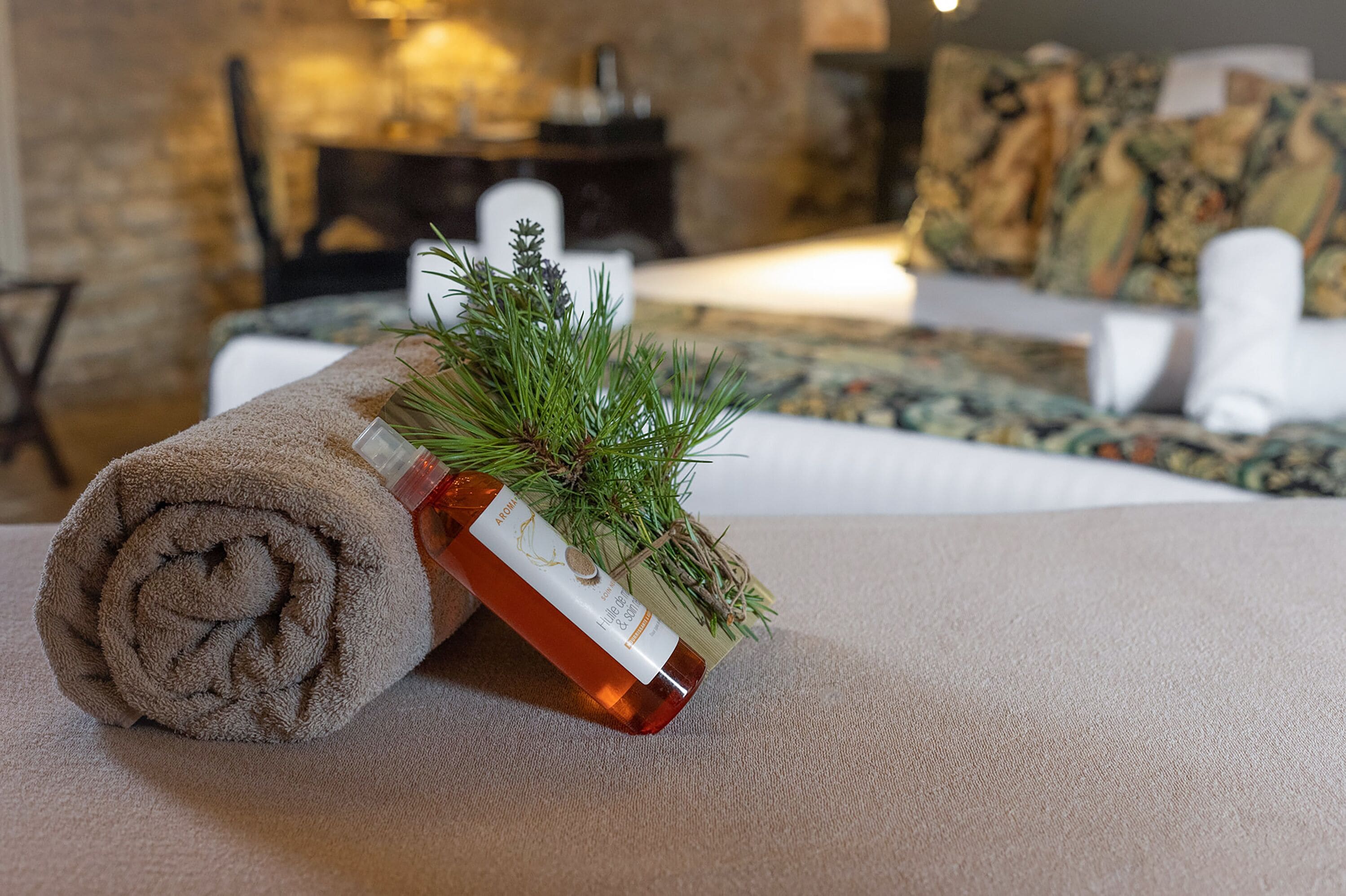 A neatly rolled brown towel on a textured surface, a bottle with a label, and a sprig of greenery.