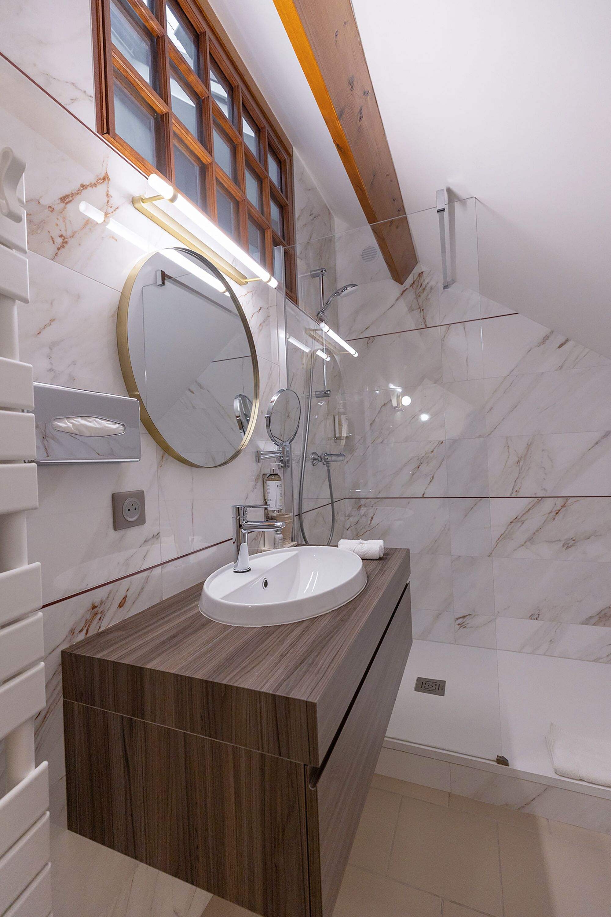 A modern bathroom with a marble wall, a wooden vanity, a round mirror, and a glass-enclosed shower.