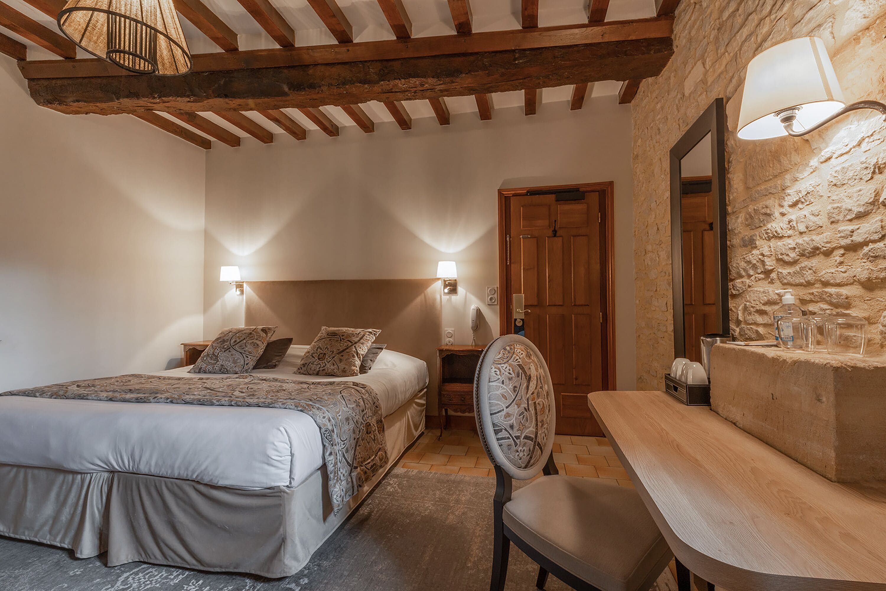 Een slaapkamer met een bed, bedlampen, een houten stoel, een tafel en een stenen muur.