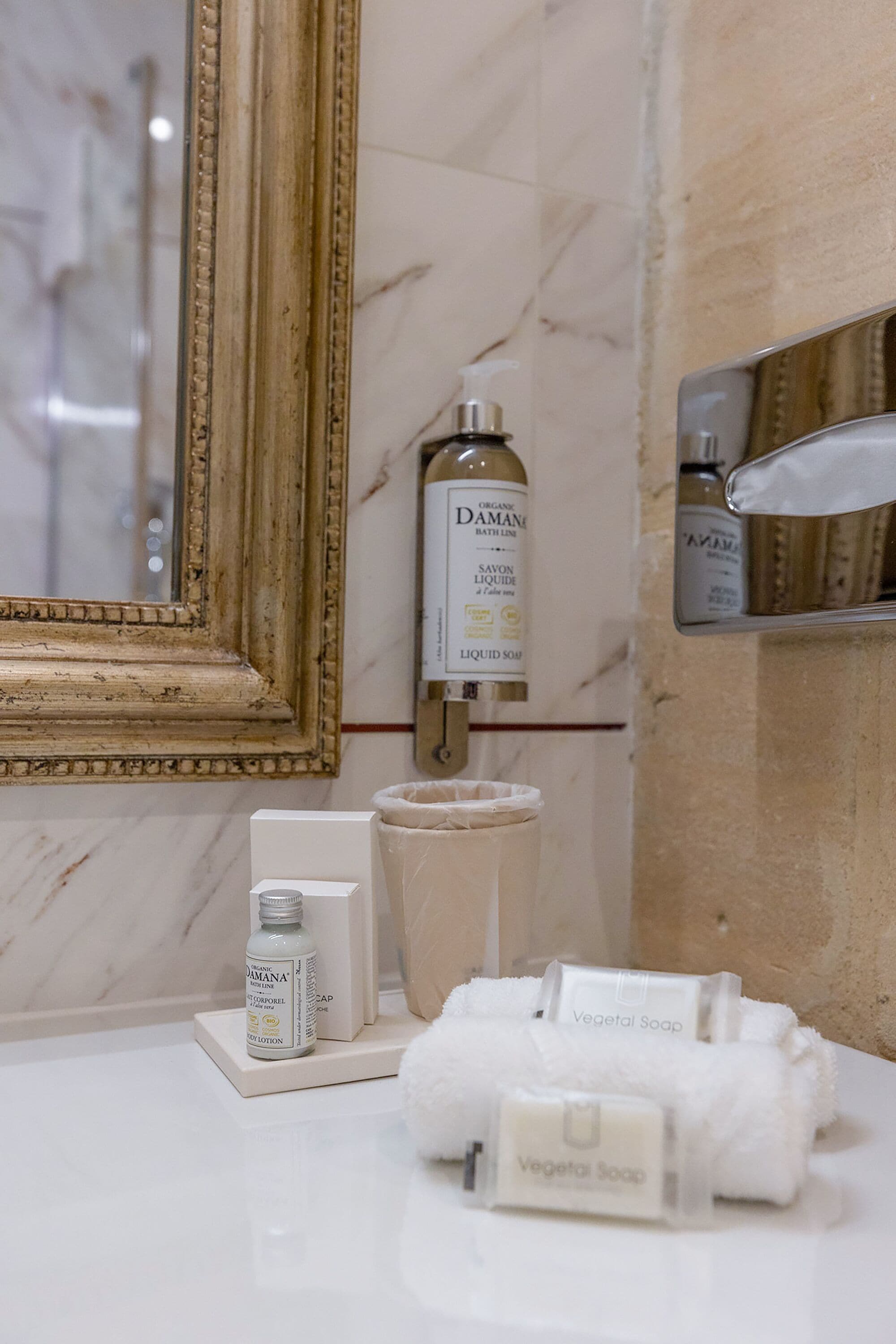 A bathroom vanity with a marble countertop, a gold-framed mirror, and toiletries including a bottle of Damani soap and a bar of Vegetal Soap.