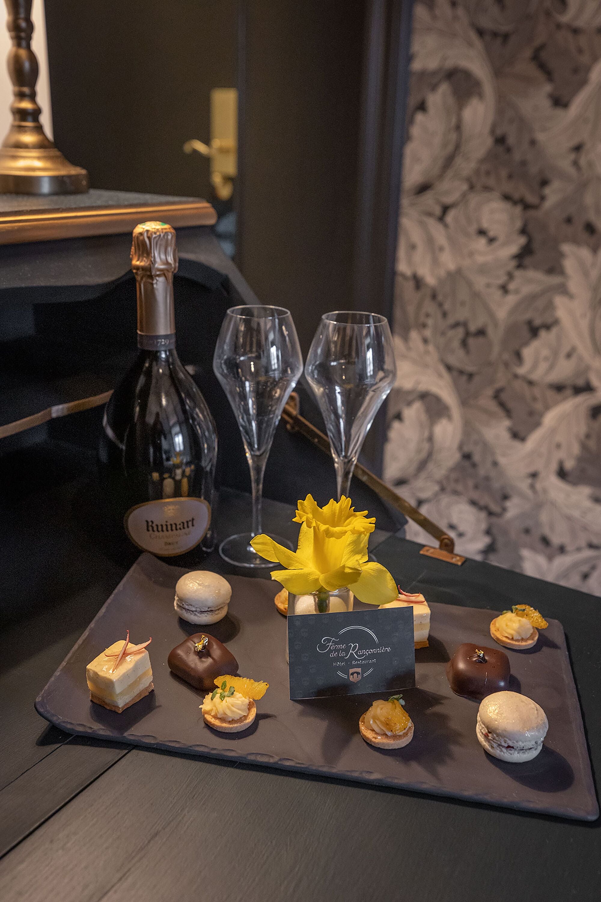 Eine Dessertplatte mit verschiedenen Süßigkeiten und einer Flasche Ruinart Wein, begleitet von zwei Sektgläsern, auf einem dunklen Tisch mit einem Hintergrund von gemustertem Wandpapier.