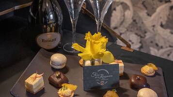 Eine Dessertplatte mit verschiedenen Süßigkeiten und einer Flasche Ruinart Wein, begleitet von zwei Sektgläsern, auf einem dunklen Tisch mit einem Hintergrund von gemustertem Wandpapier.