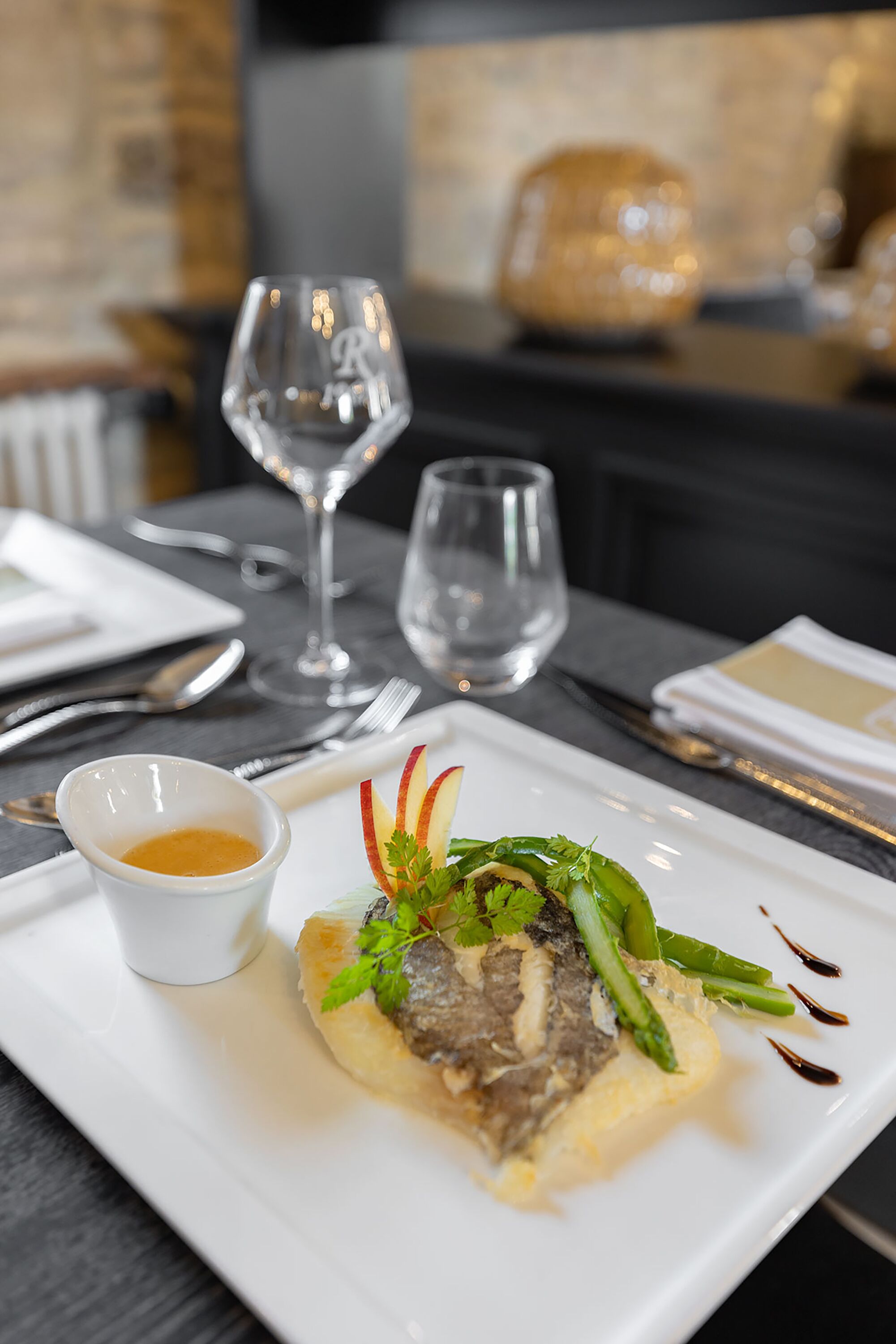 A white plate with a steak, asparagus, and greens, accompanied by a small bowl of sauce, on a table with cutlery and glasses.