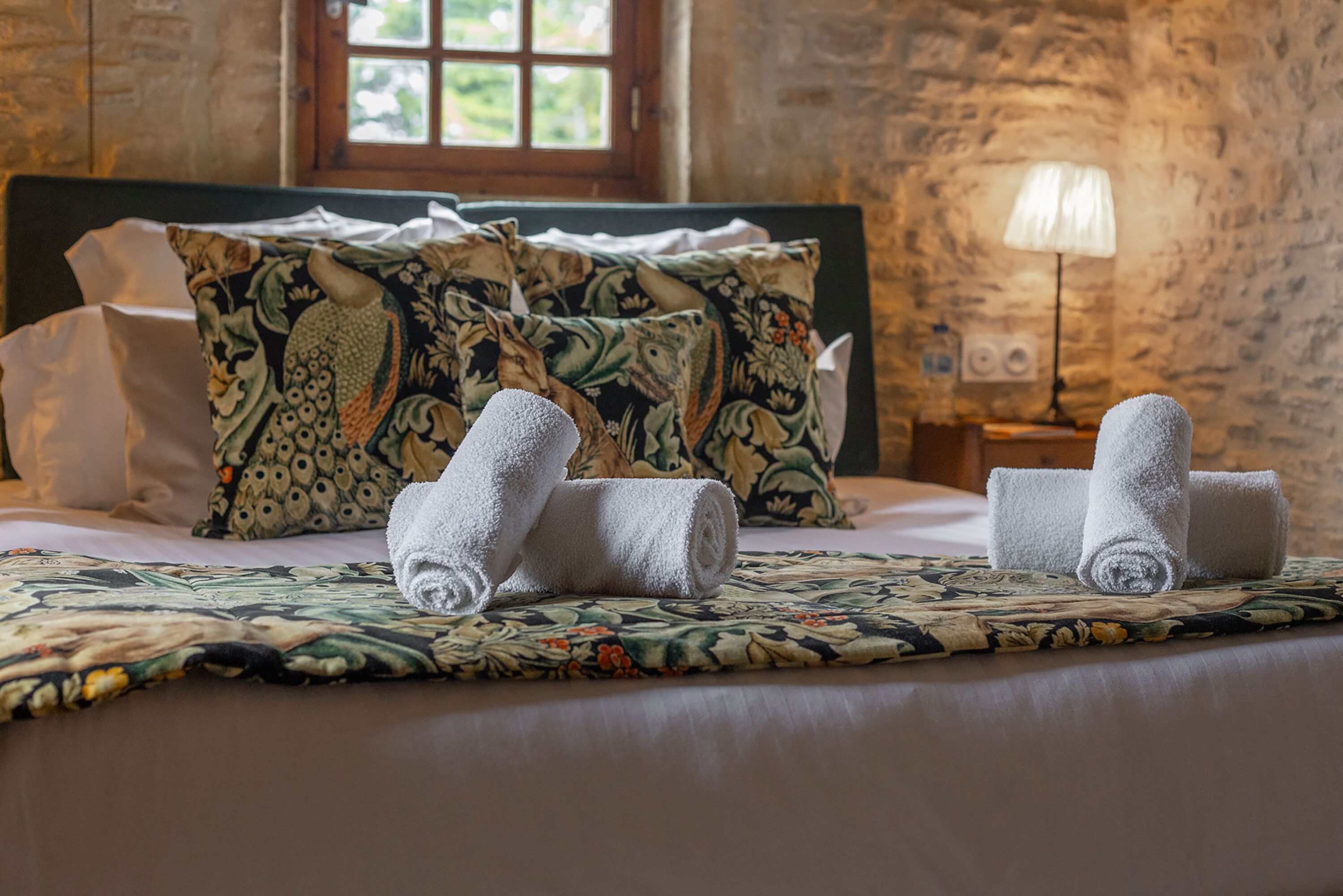 A neatly made bed with floral-patterned pillows and rolled towels, a lamp on a bedside table, and a stone wall in the background.