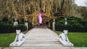 A wooden pathway leading to a structure under a weeping willow tree, with decorative lights and statues on either side.