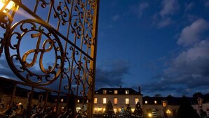 A wrought iron gate with a large 'H' emblem, a building with lit windows, and Christmas trees in the foreground.