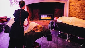 A spa room with a stone wall, a fireplace, and two massage tables.