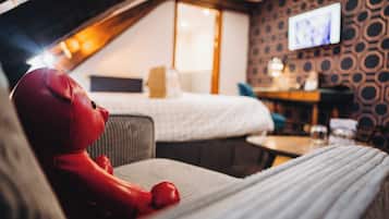 Habitación de hotel con cama, televisor montado en la pared, una mesita y un peluche rojo sobre la cama.