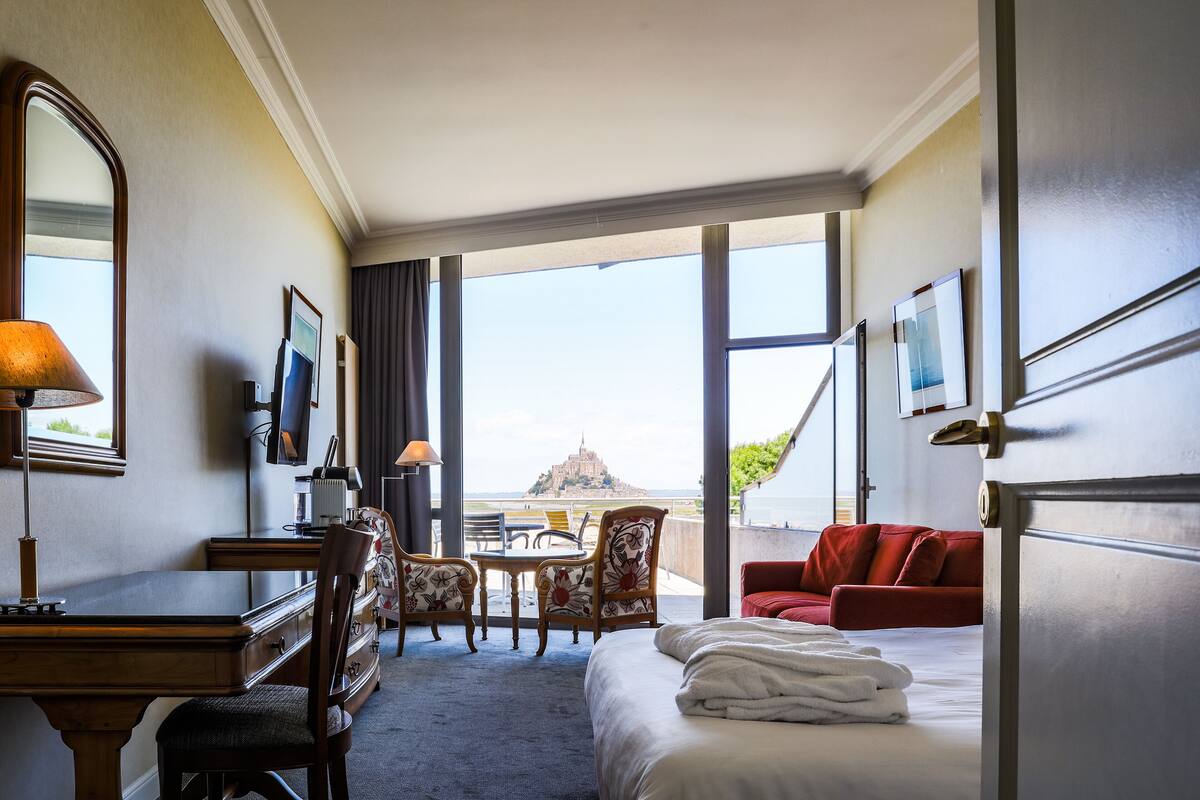 A hotel room with a bed, desk, chairs, and a view of a distant landmark.