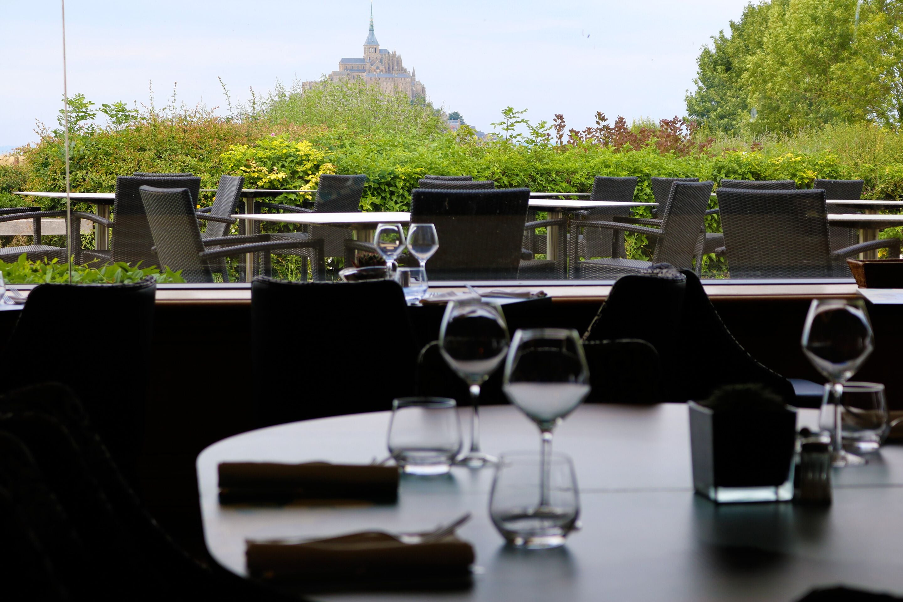 An outdoor dining area with tables and chairs, a view of a castle in the background, and a lush green garden.