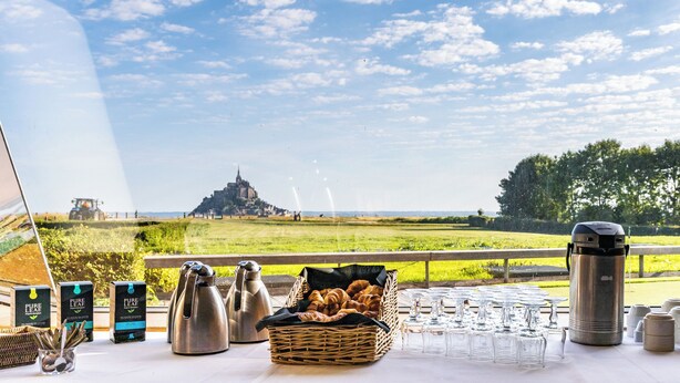 A table set with breakfast items, a thermos, and glasses, overlooking a scenic landscape with a distant castle.