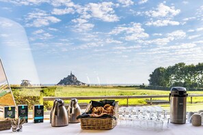 A table set with breakfast items, a thermos, and glasses, overlooking a scenic landscape with a distant castle.