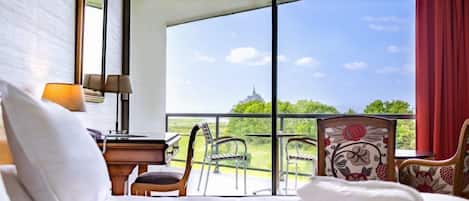 A hotel room with a bed, a desk, a chair, and a balcony with a view of greenery and a distant building.