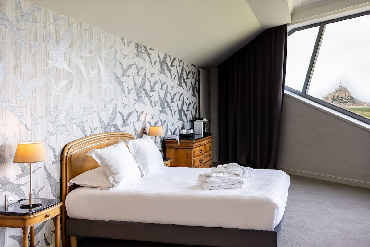 A hotel room with a bed, a nightstand, a lamp, a dresser, and a window with a view of a distant building.