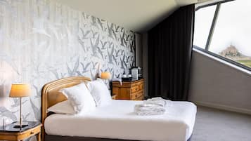 A hotel room with a bed, a nightstand, a lamp, a dresser, and a window with a view of a distant building.