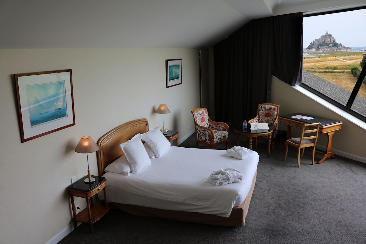 A hotel room with a bed, a desk, a chair, a lamp, and a view of a castle.