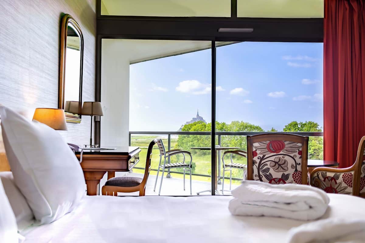 A hotel room with a large bed, a desk with a lamp, and a view of a balcony with chairs and a table.