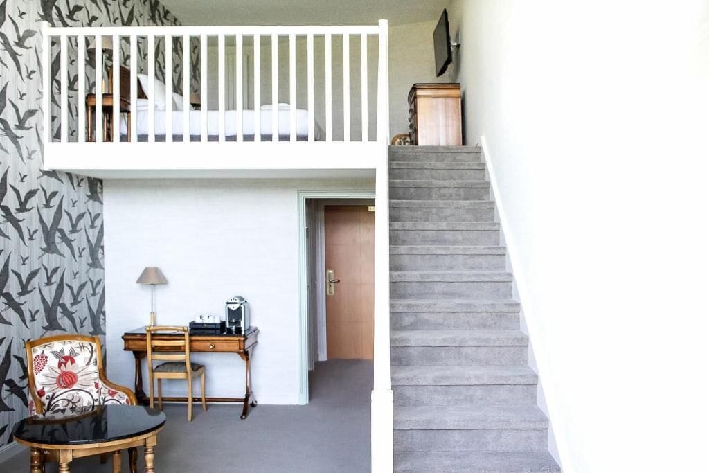 A room with a bunk bed, a desk with a lamp, a chair, and a staircase.