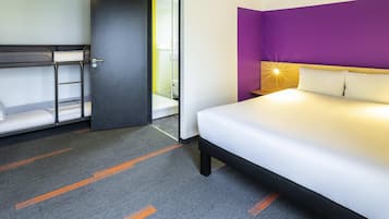A hotel room with a bunk bed, a single bed, a purple accent wall, and a nightstand with a lamp.