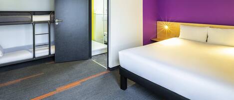 Una habitación de hotel con una cama litera, una cama individual, una pared de acento morada y una mesita de noche con lámpara.