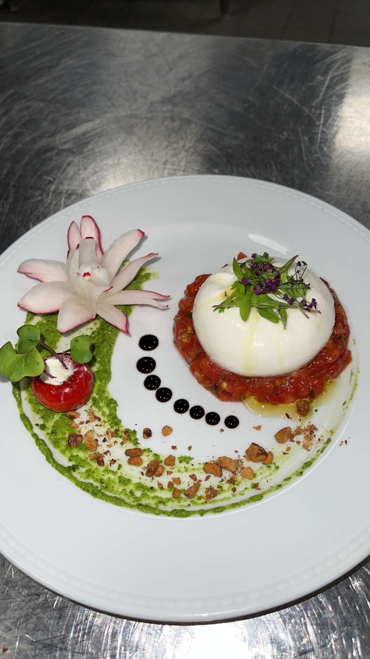 A white plate with a round white food item, green sauce, black olives, and a cherry tomato.