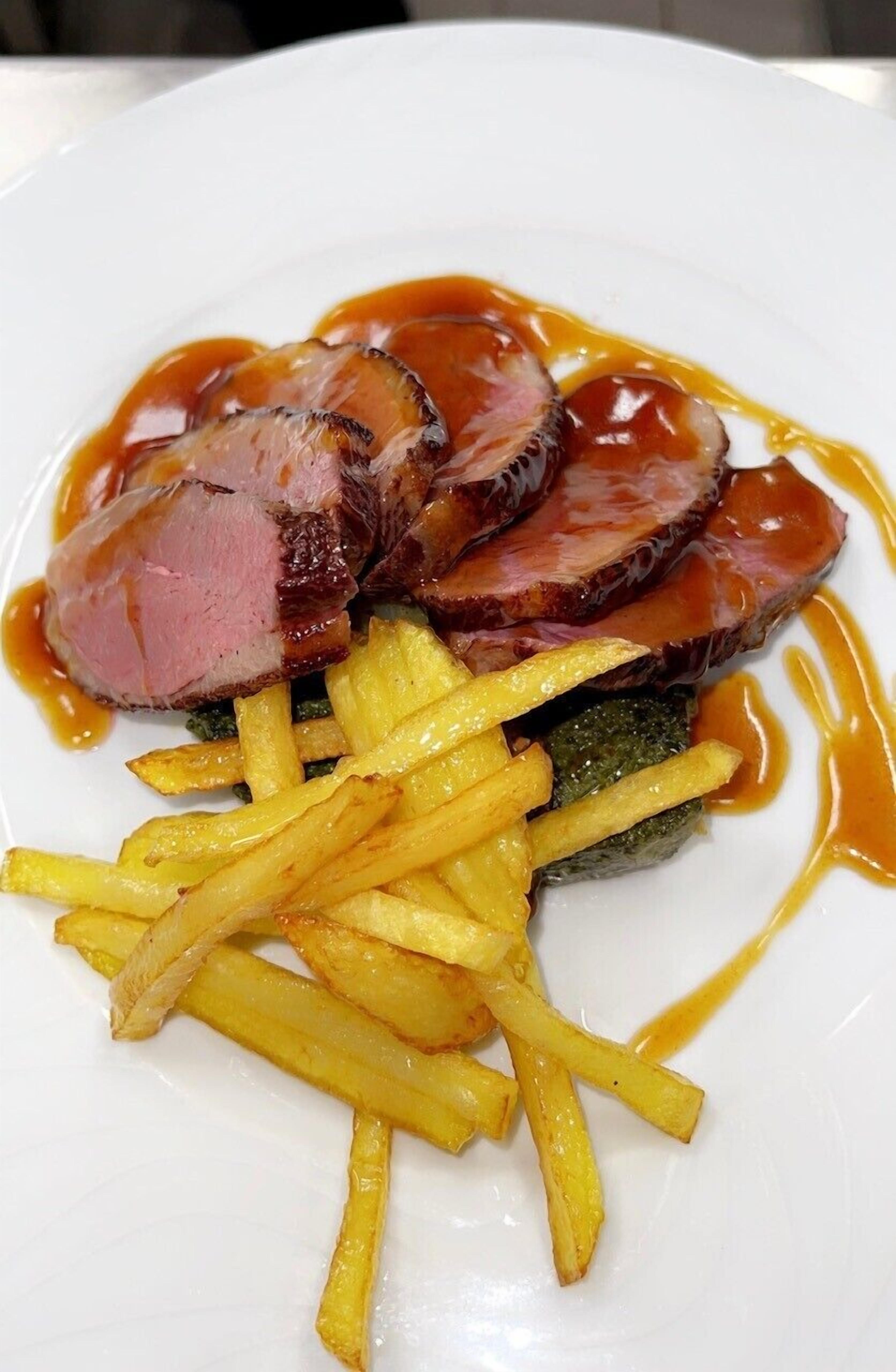 A plate with slices of meat, fries, and a green vegetable, possibly broccoli, with a sauce.