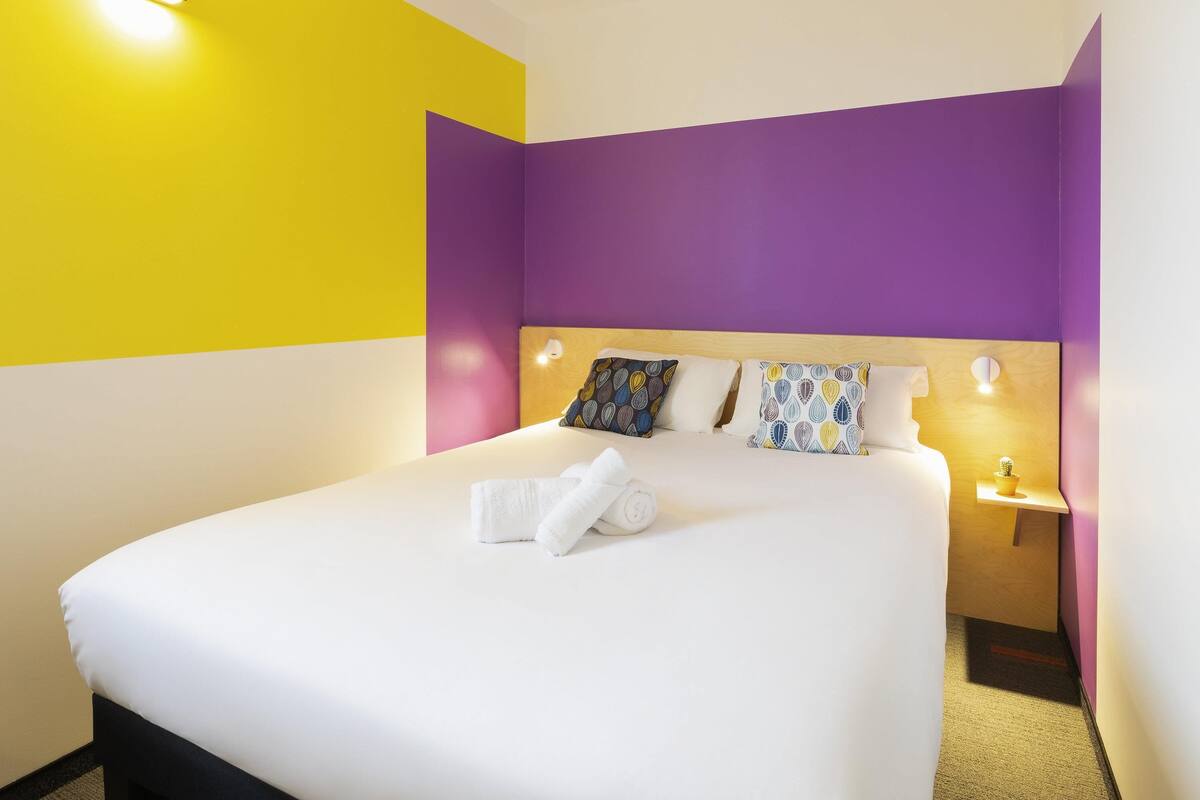 Una habitación de hotel con una cama, una mesita de noche y una lámpara de pared.