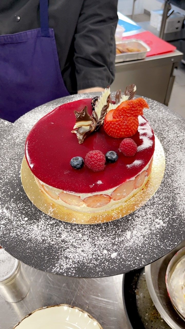 A round dessert with a glossy red top, strawberries, and blueberries on a cake stand.