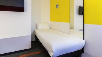 A single bed in a room with a yellow accent wall, a flat-screen TV mounted on the wall, and a small table with a chair.