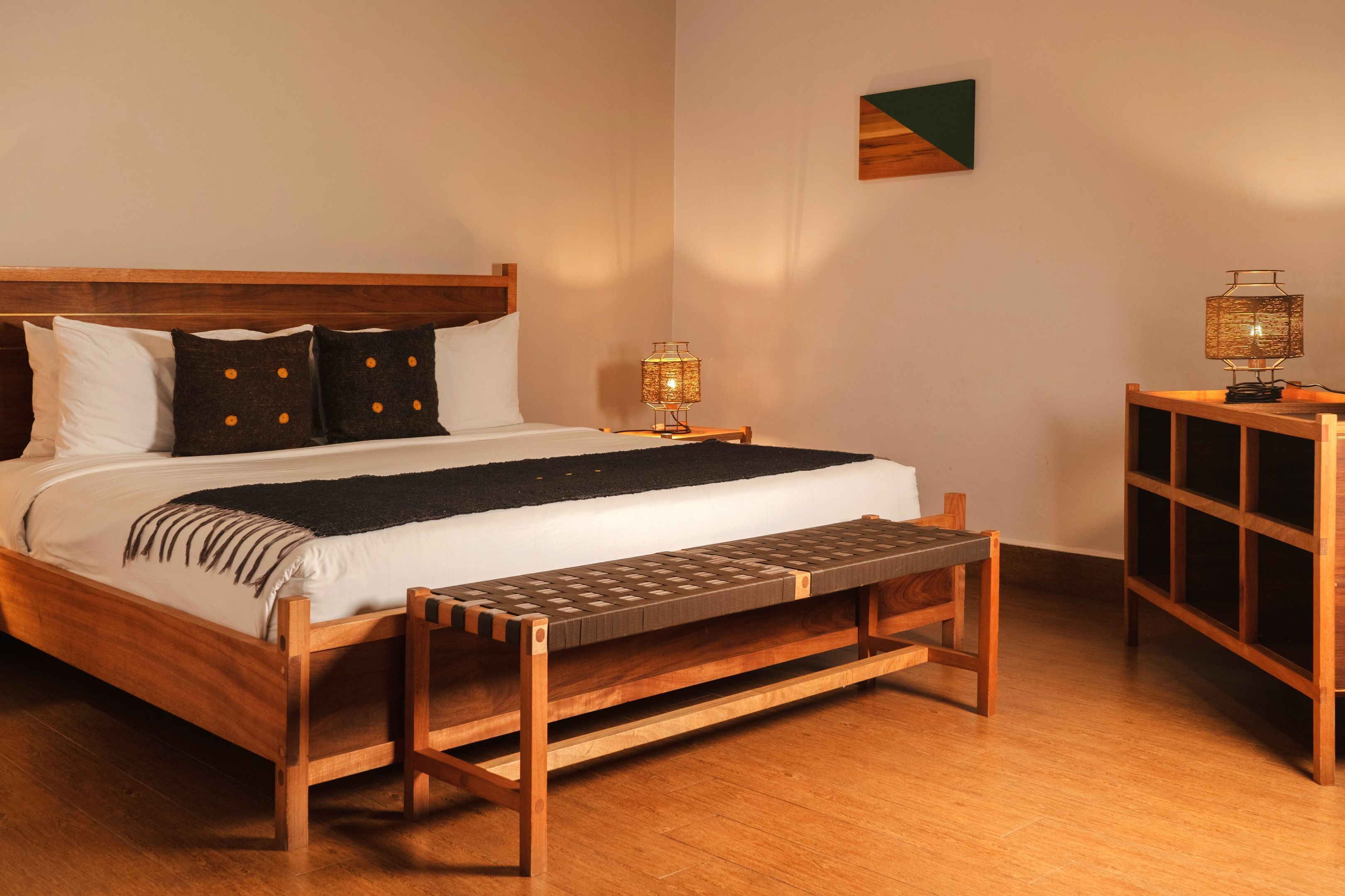 Una cama bien hecha con cabecera de madera, un banco al pie de la cama, una mesita de noche con lámpara y un mueble pequeño.