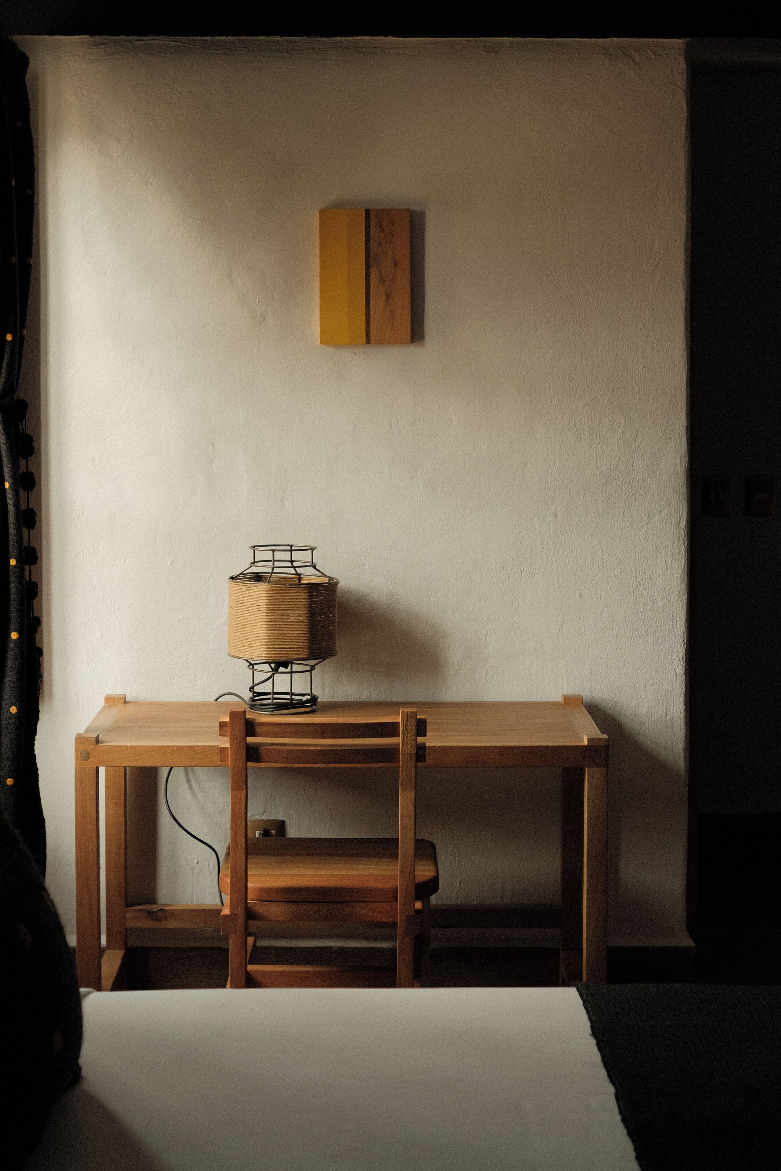 Un escritorio de madera con una lámpara, una silla y una mesita auxiliar frente a una pared lisa.