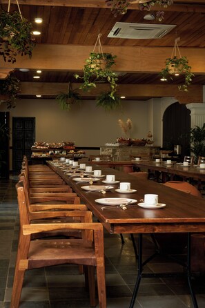 A long wooden dining table set for a meal with chairs, hanging plants, and a kitchen area in the background. - Hotel Sombra del Agua (San Cristóbal de las Casas)