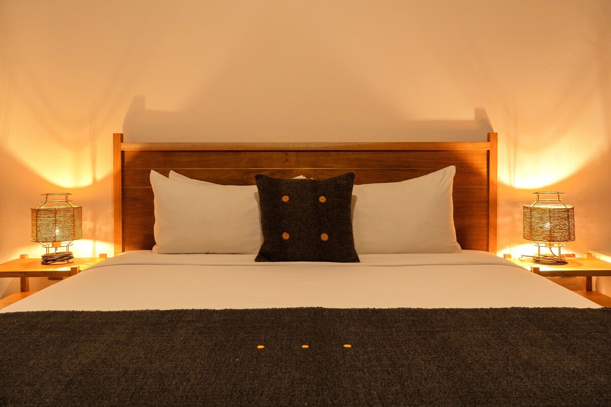 A neatly made bed with a wooden headboard, white pillows, and a dark throw blanket. There are two bedside lamps with patterned shades on either side of the bed.