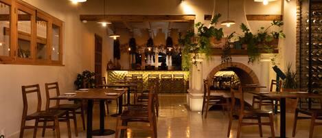 Interior de restaurante con mesas y sillas de madera, luces colgantes y plantas en macetas.