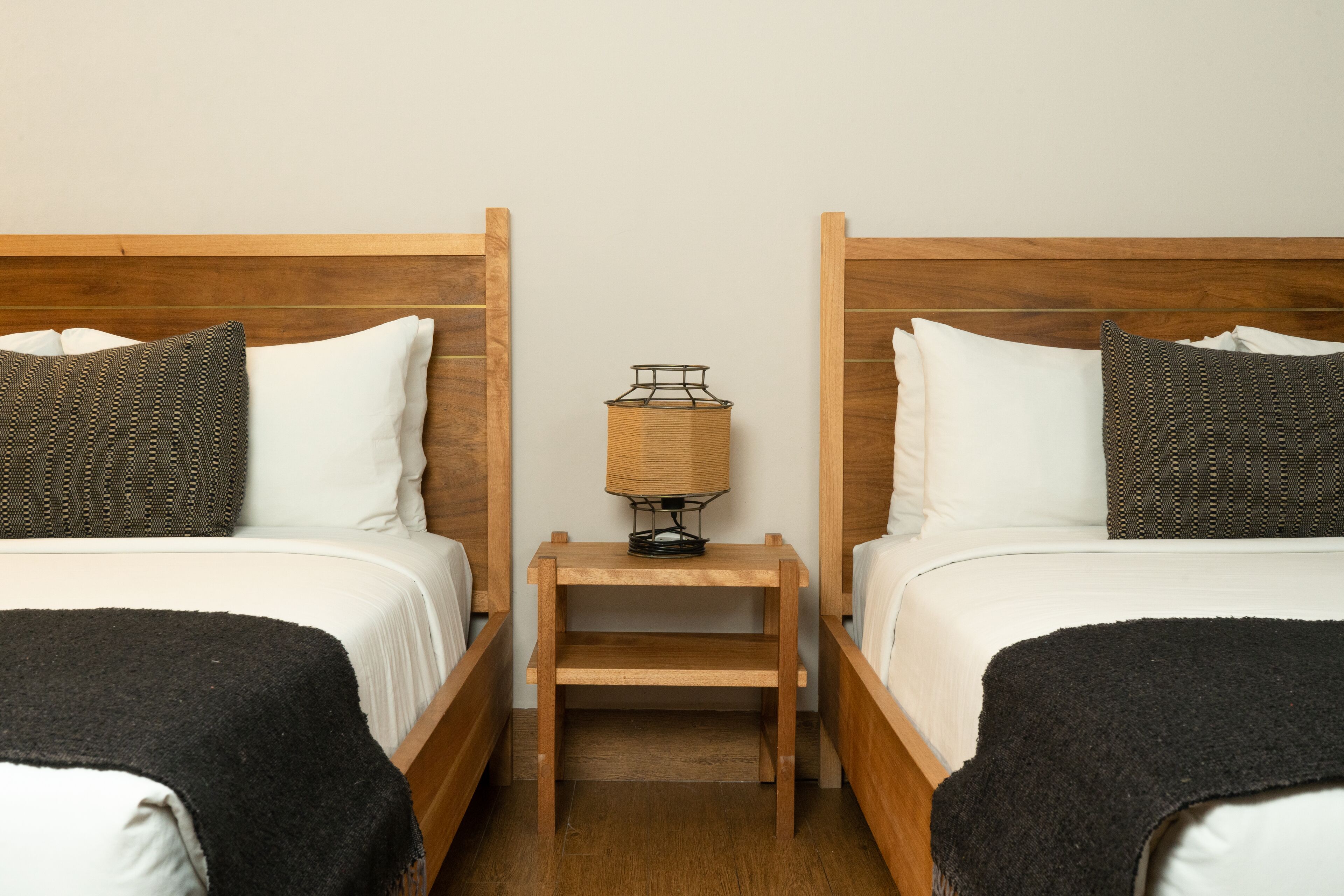 Habitación doble con dos camas, cada una con cabecero de madera, una mesita de noche de madera con lámpara y una manta de color negro.