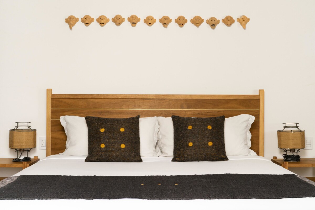 A neatly made bed with a wooden headboard, two white pillows, two brown pillows with yellow buttons, and a black throw blanket. There are two bedside lamps with wicker shades on wooden nightstands.