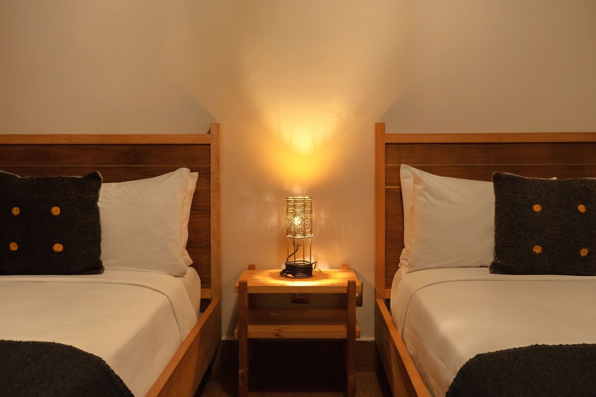 A hotel room with two wooden beds, each with white bedding and dark pillows with yellow buttons. A small wooden nightstand with a lamp is between the beds.