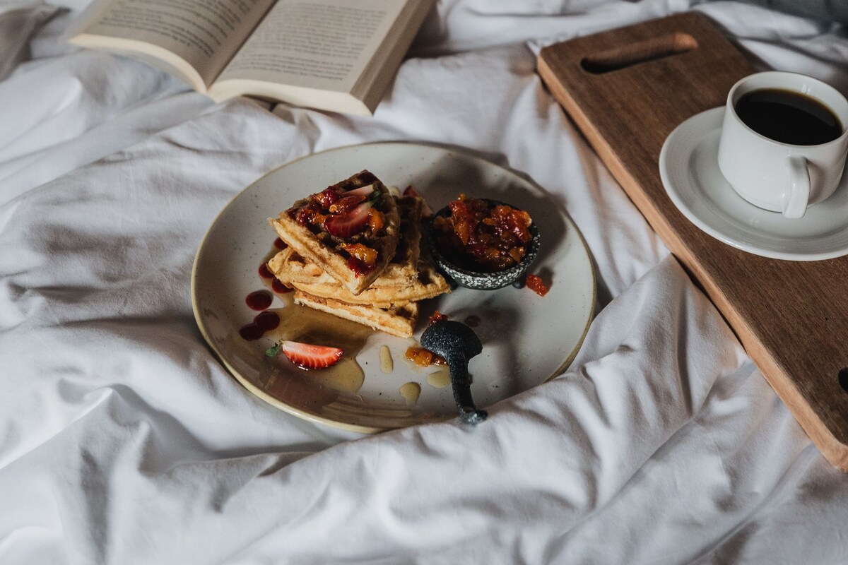 A bed with a book, a plate of food, a cup of coffee, and a tray.