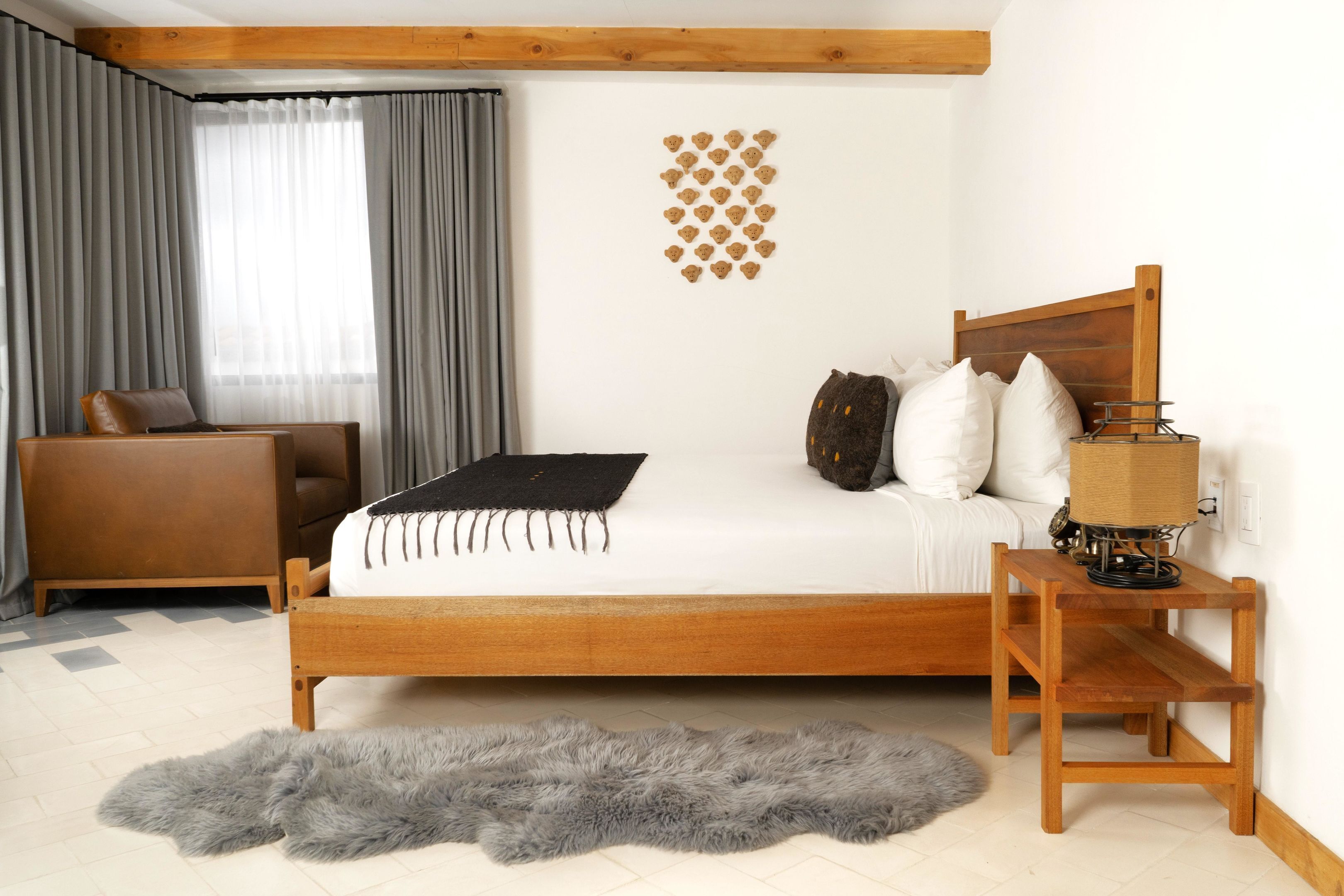 A bedroom with a wooden bed, a small wooden nightstand, a grey furry rug, a brown leather chair, and a window with grey curtains.
