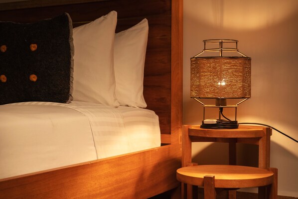 A bed with white linens and a dark pillow, a wooden nightstand with a woven lamp, and a small wooden chair.