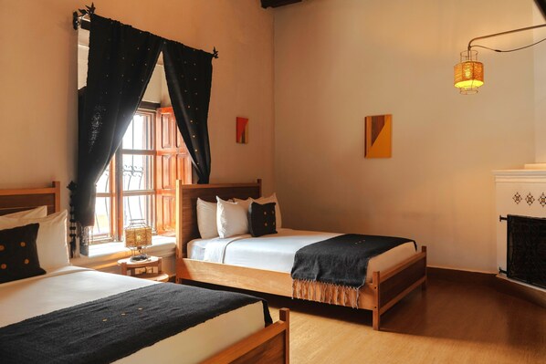 A room with two beds, a window with curtains, a bedside table with a lamp, and a wall-mounted light fixture.