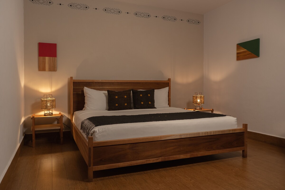 A bedroom with a wooden bed, two bedside tables with lamps, a red and green wall decoration, and a patterned ceiling.